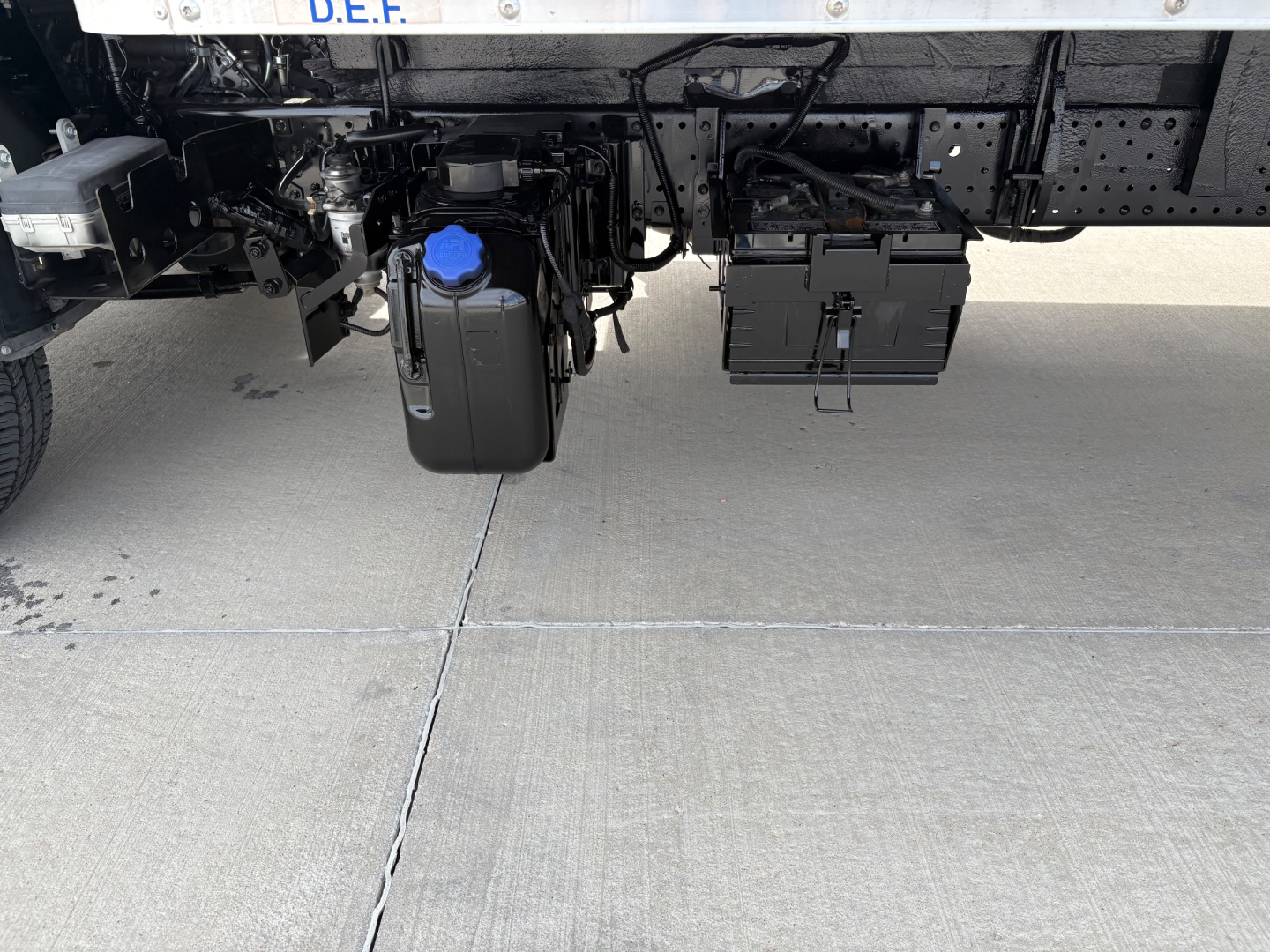 Underbody view of a 2019 Isuzu NPR-HD truck showing a blue-capped windshield washer reservoir and a black battery box mounted under the frame.