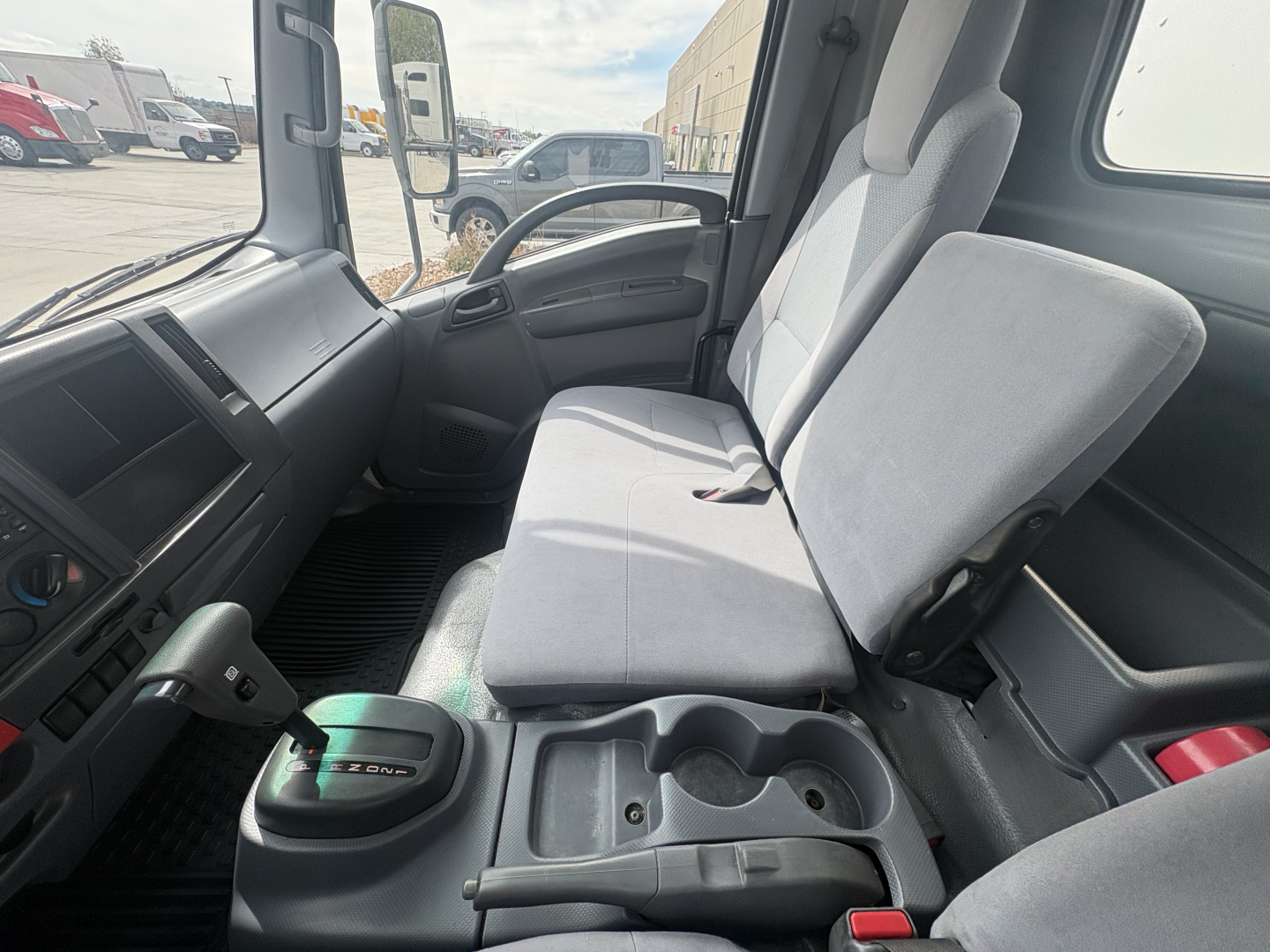 Isuzu NPR-HD cab interior showing front bench seat and center console with cup holders and gear selector; steering wheel and dashboard visible.