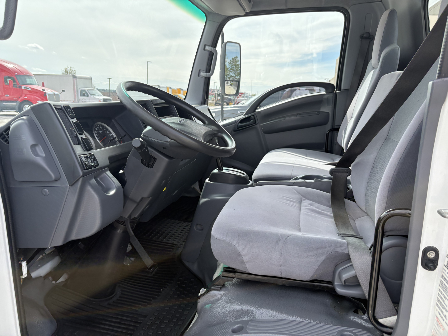 Isuzu NPR-HD cab interior showing steering wheel, driver seat, and dashboard with gray fabric seats and open door.