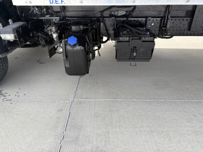 Underbody view of a 2019 Isuzu NPR-HD truck showing a blue-capped windshield washer reservoir and a black battery box mounted under the frame.