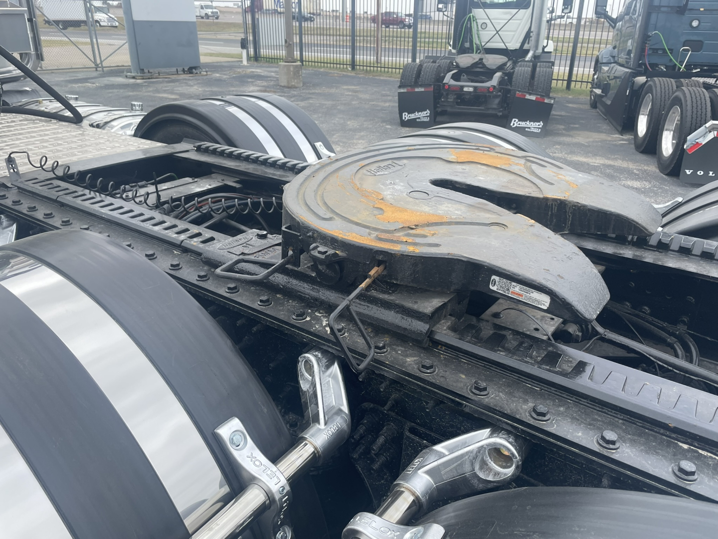 Truck chassis with fifth wheel and fuel tank area. Visible components include a large dual-rail fifth wheel coupling plate with rust patches, air lines, and a shiny chrome pin or hinge. No readable labels or text clearly visible in this vie