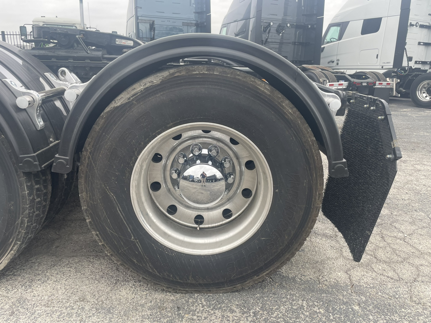 Front wheel and fender area of a new Mack GR84FT conventional daycab showing large front wheel with hub and chrome center, mudguard, and wheel well.