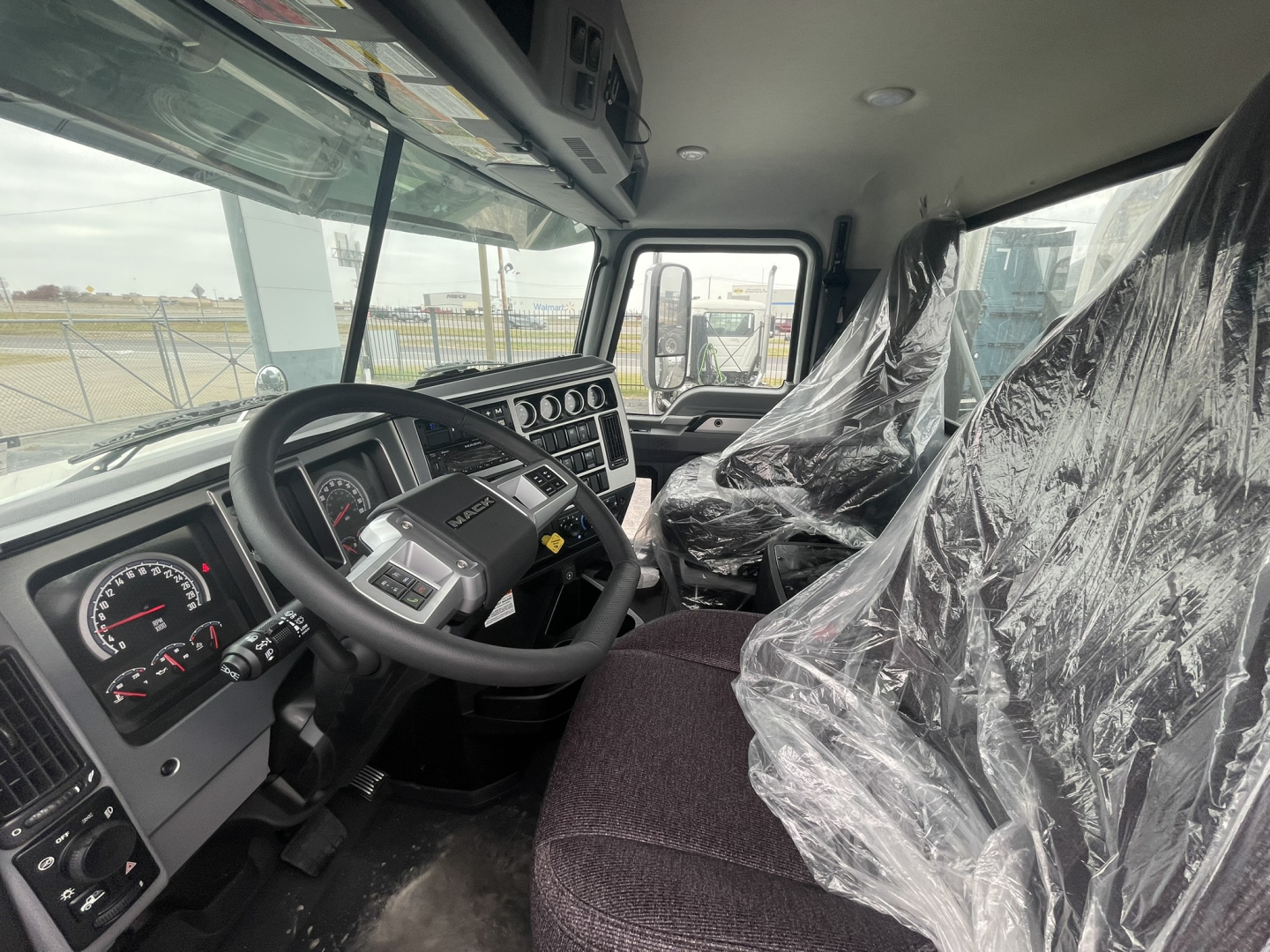 Truck cab interior showing steering wheel, instrument cluster, and driver and passenger seats covered in plastic. Dashboard with gauges and controls visible.