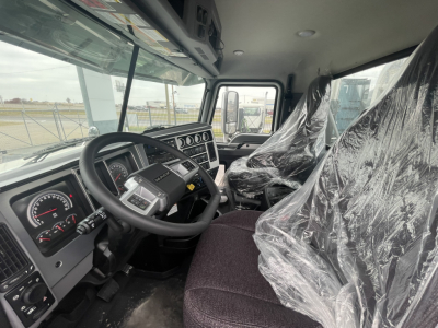 Truck cab interior showing steering wheel, instrument cluster, and driver and passenger seats covered in plastic. Dashboard with gauges and controls visible.