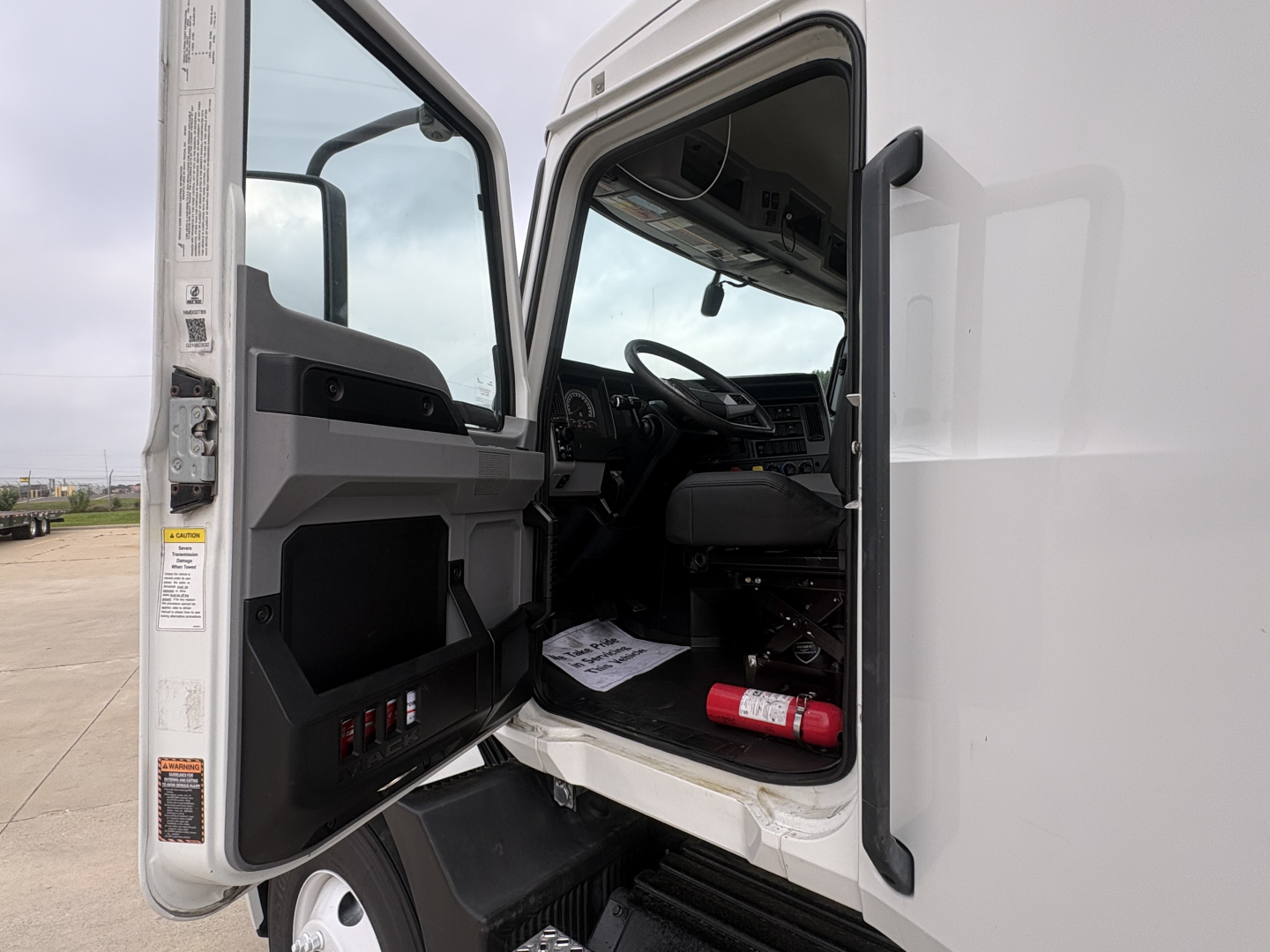 Truck cab interior with open door showing steering wheel and dashboard; fire extinguisher mounted near floor; gray interior panels; window and mirror visible.