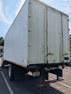 Back view of a white box truck cargo area with rear doors and bumper, showing a California license plate and dual rear wheels.