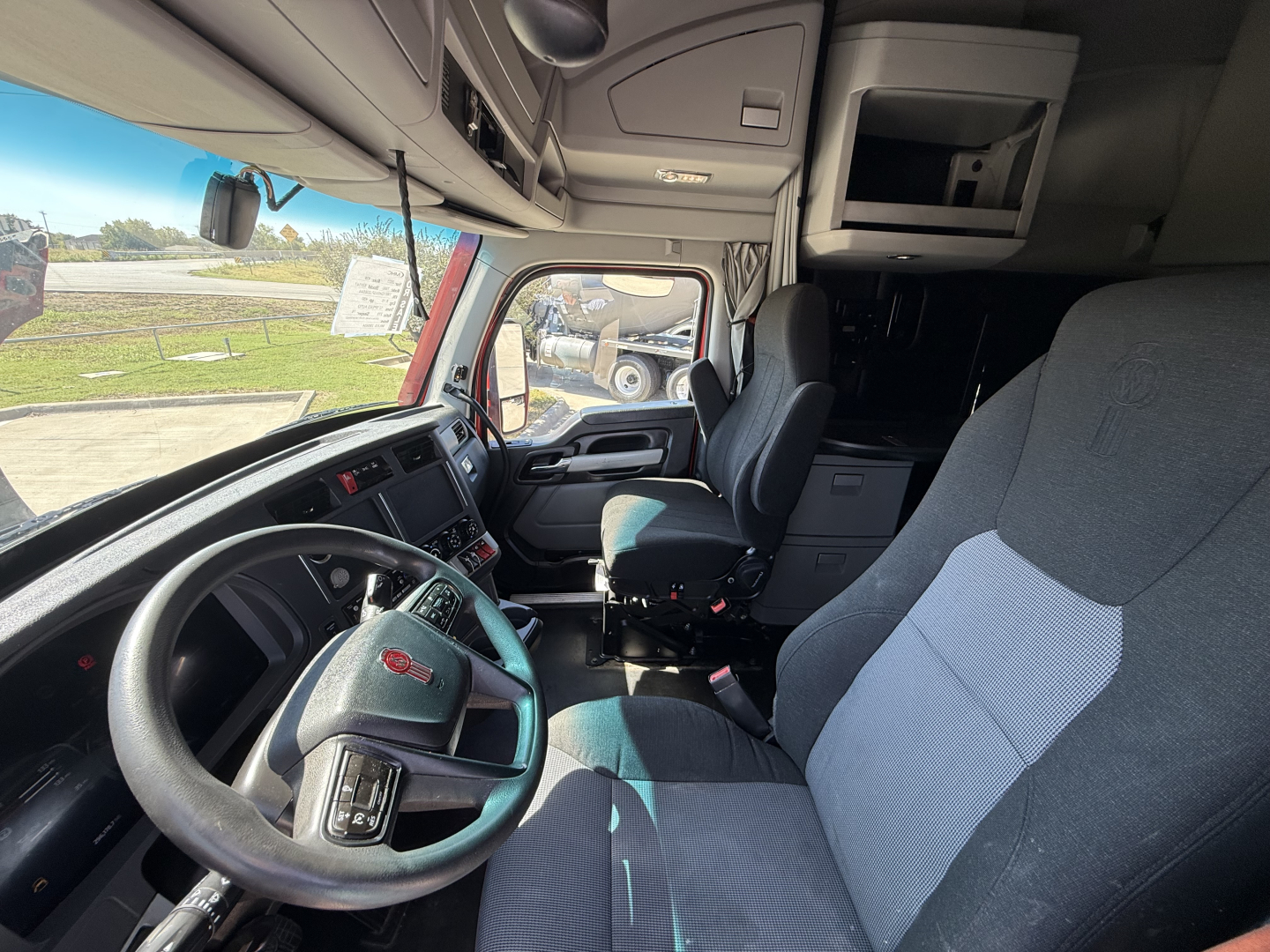 Kenworth T680 truck cab interior view showing steering wheel, driver seat, passenger seat, dashboard controls, overhead storage, and window; part of a used truck listing.