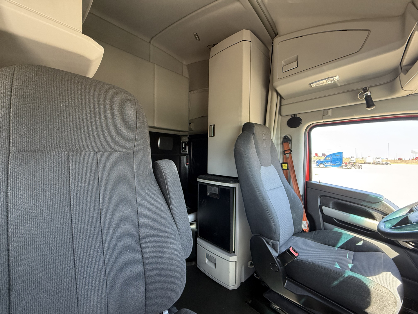 Kenworth T680 cabin interior showing driver seat and sleeper cab with overhead storage and small fridge unit.