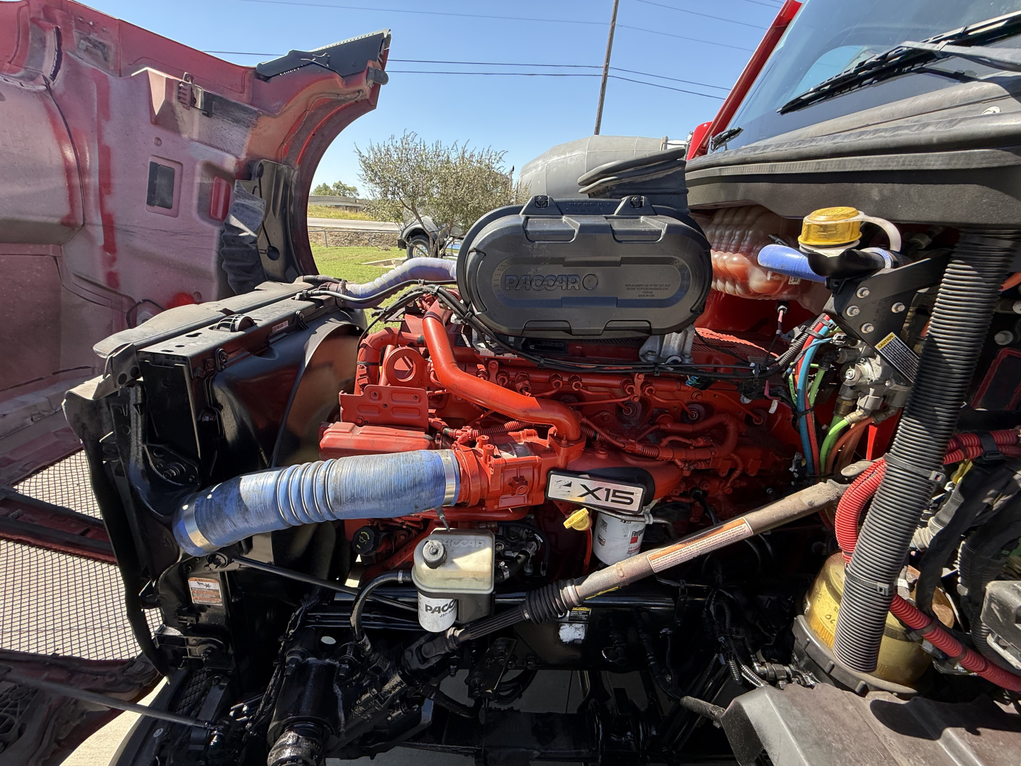 Kenworth T680 engine bay with bright orange engine block, blue intake hose, and exposed components under open hood.