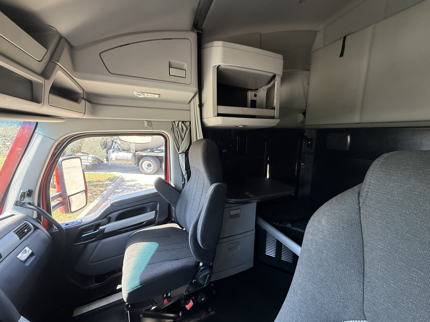 Truck cab interior of a Kenworth T680 with driver seat, upper storage cabinet, desk area, and sleeping berth visible.