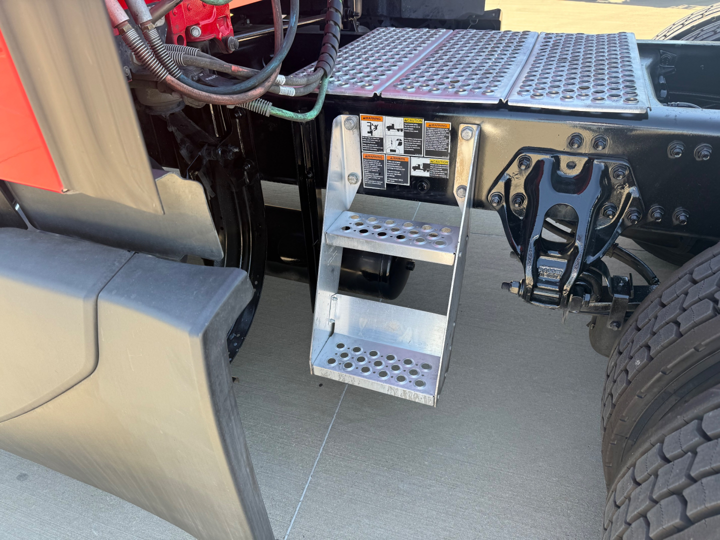 Kenworth T680 cab area showing metal step/ladder with perforated steps and sticker labels near chassis, front wheel, and wiring harness.