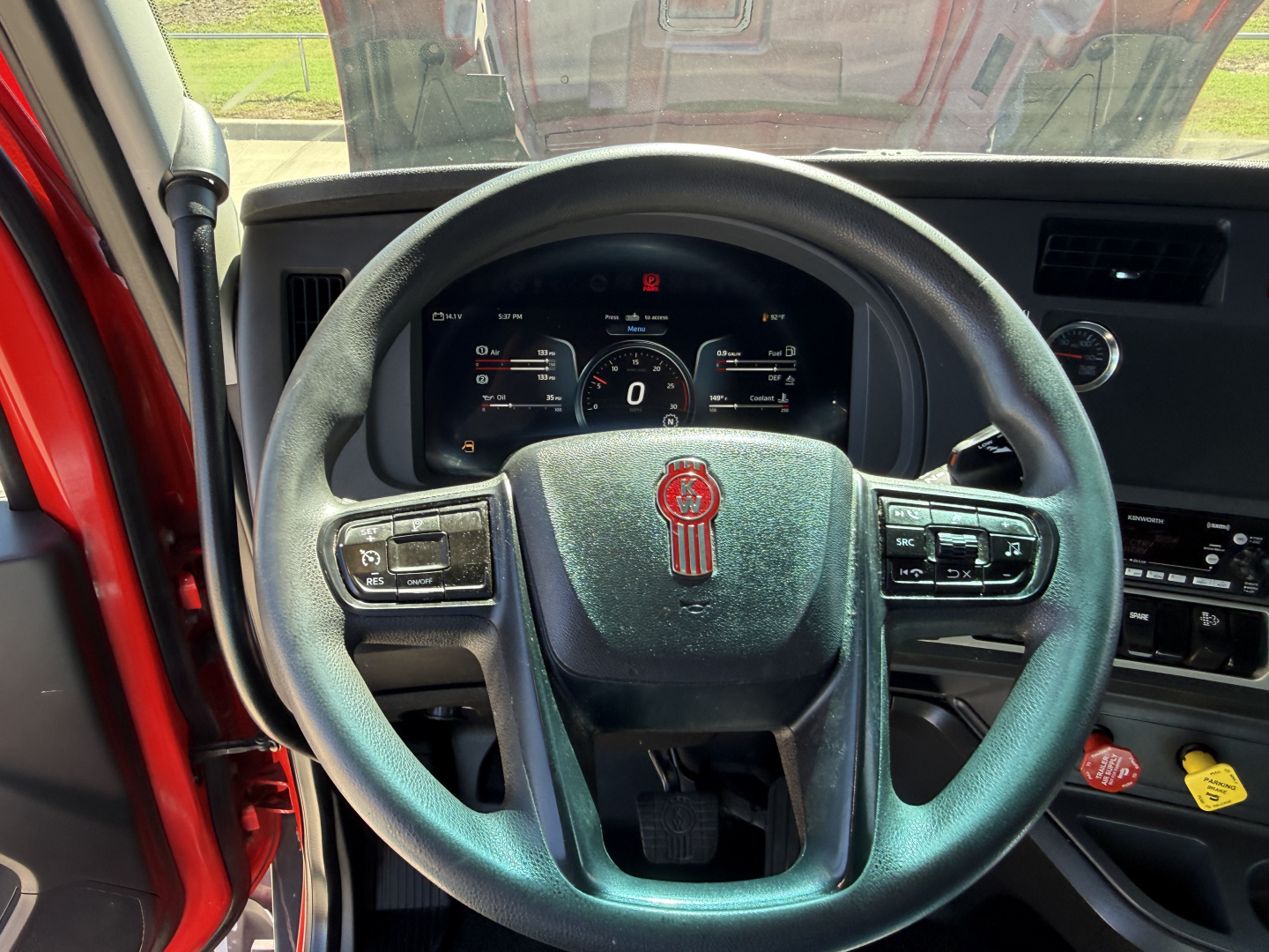 Kenworth T680 steering wheel and dashboard view with digital instrument cluster in center, red exterior visible through open driver door.