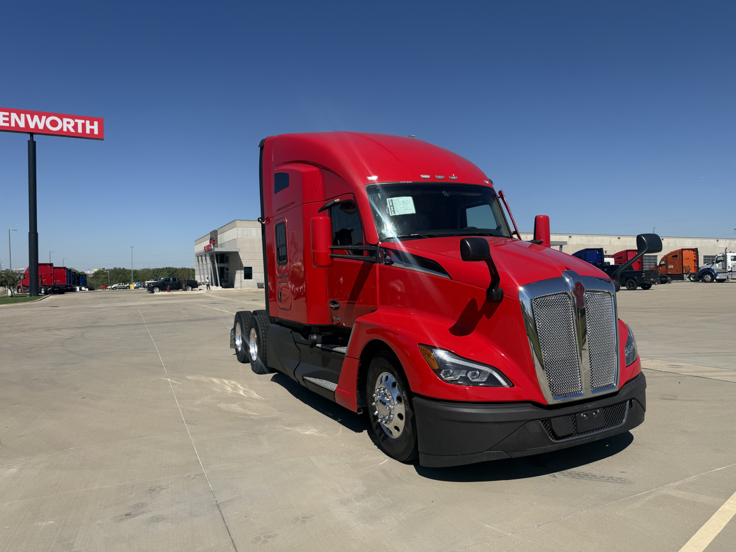 Kenworth T680 truck in red cab with sleeper, front grille, and side mirrors visible; background shows dealership lot with other trucks and buildings.