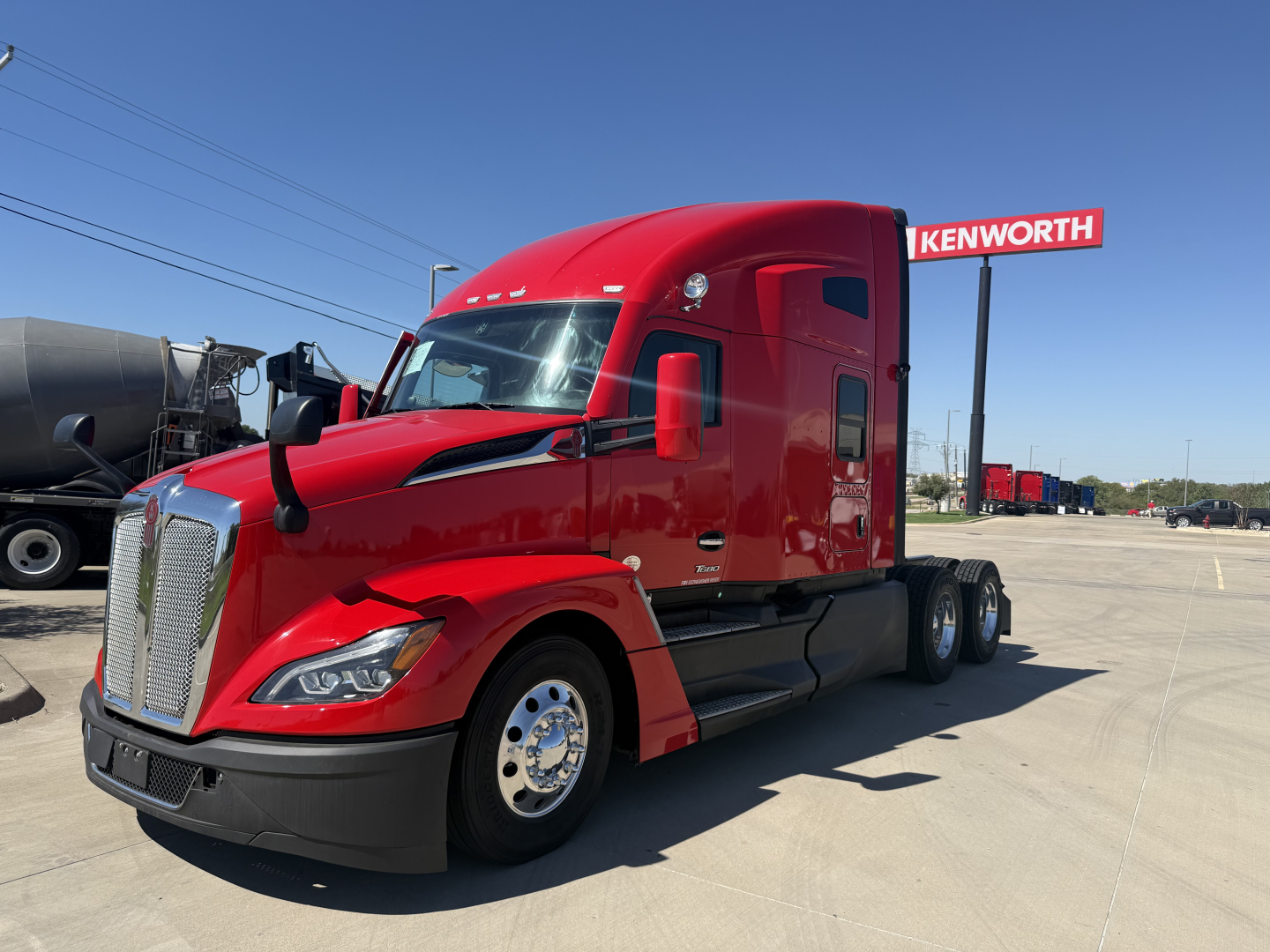 Kenworth T680 conventional sleeper truck in red, side view, showing front grille, chrome wheel covers, and Kenworth sign in background.