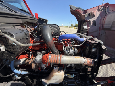 Kenworth T680 engine bay view with blue coolant hoses, red engine block, and various hoses and components visible under open hood in a truck yard
