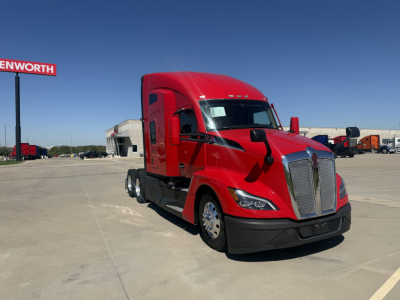 Kenworth T680 truck in red cab with sleeper, front grille, and side mirrors visible; background shows dealership lot with other trucks and buildings.