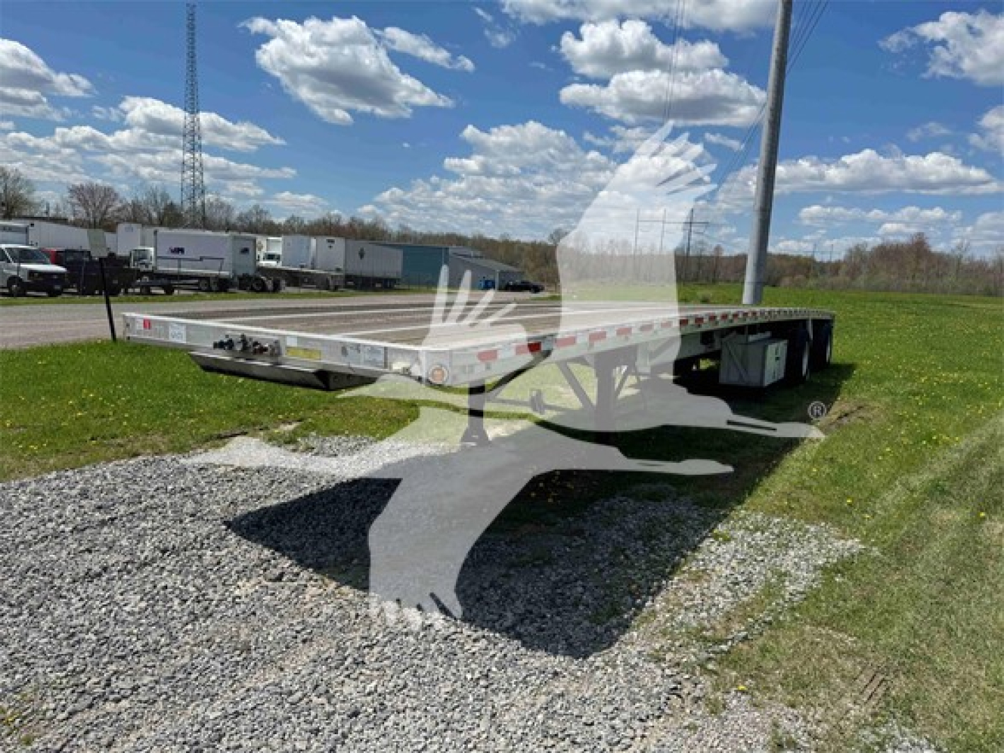 Flatbed trailer showing low-profile deck with dual axles and side rails; reflective tape along sides; stack of trailers and blue sky with clouds in background.