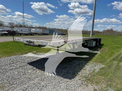 Flatbed trailer showing low-profile deck with dual axles and side rails; reflective tape along sides; stack of trailers and blue sky with clouds in background.