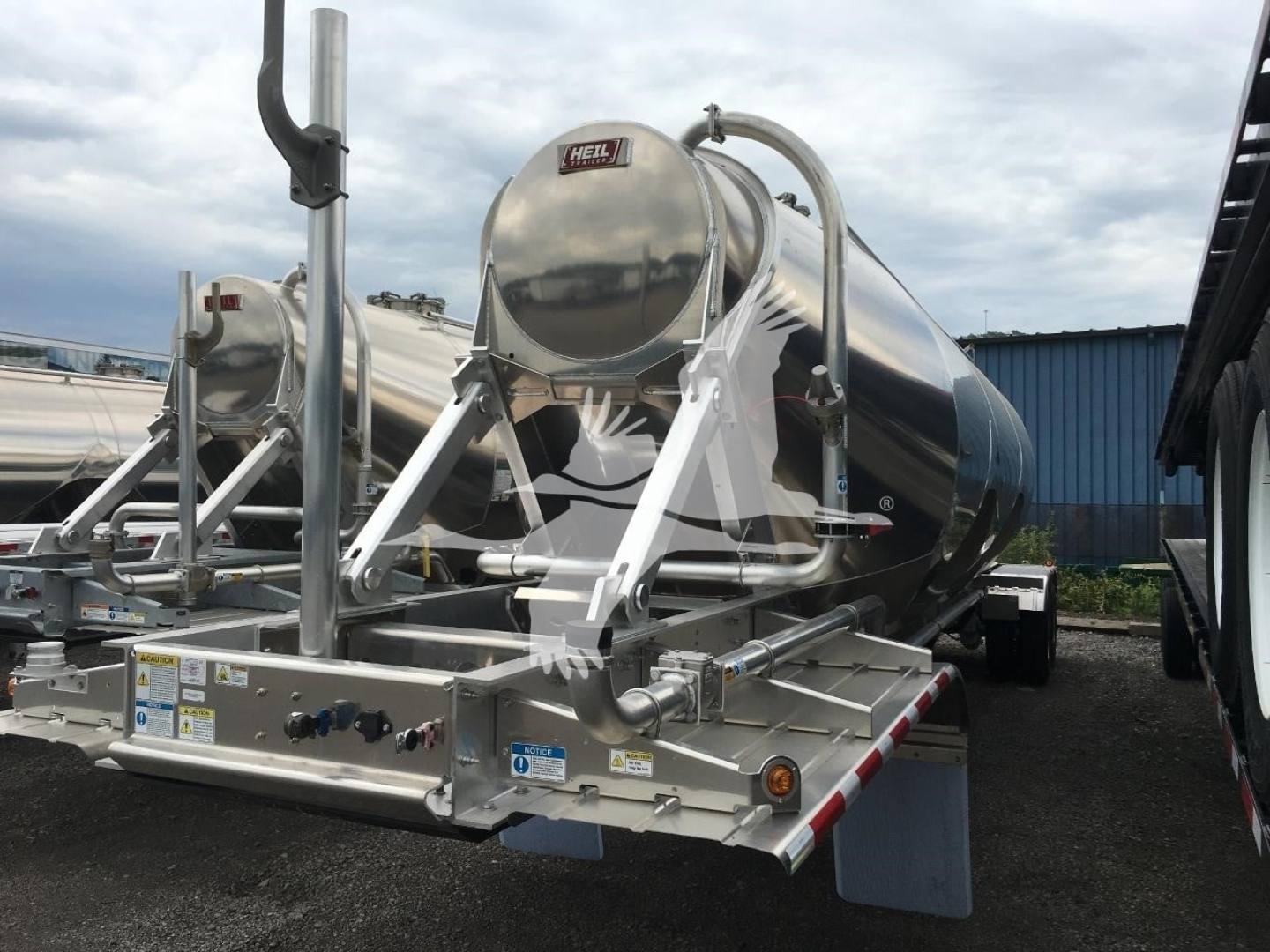 Heil cementer trailer with polished stainless steel cylindrical tank, pipes, and control panel; visible labels and safety decals on chassis.