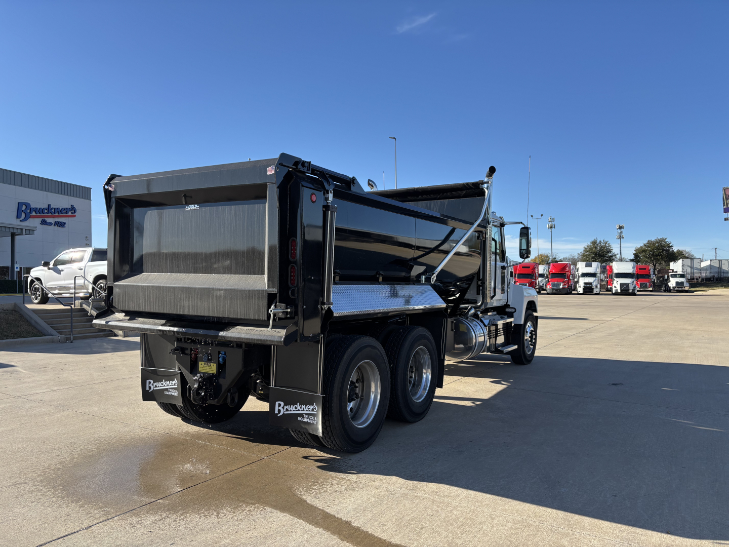 Dump truck in a dealership lot showing a new 2026 Mack PI64 dump truck with a black dump bed and dual rear axles.