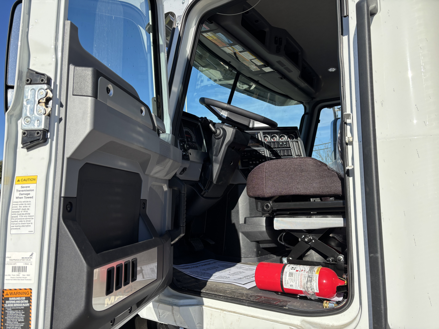 Mack PI64 truck cab interior with steering wheel, driver's seat, and dashboard; fire extinguisher on floor; door frame and warning labels visible.