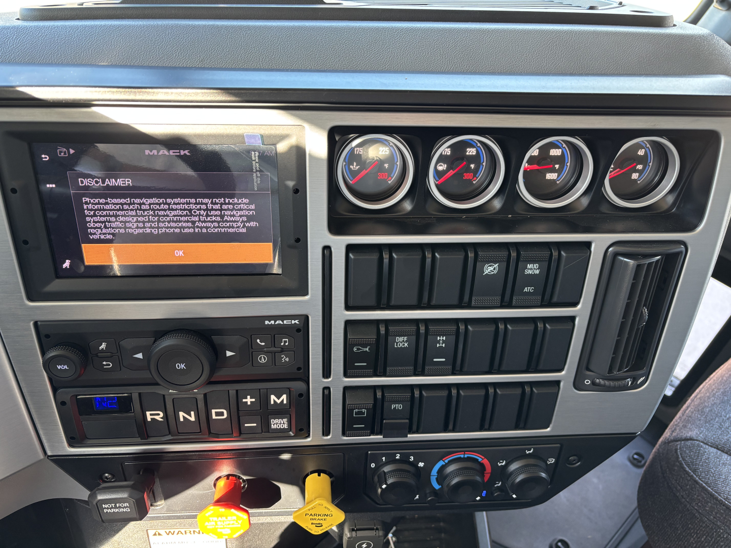 Dashboard of a 2026 Mack PI64 showing digital display, 4 round gauges, gear selector with R N D P, and control panels with buttons and climate controls.
