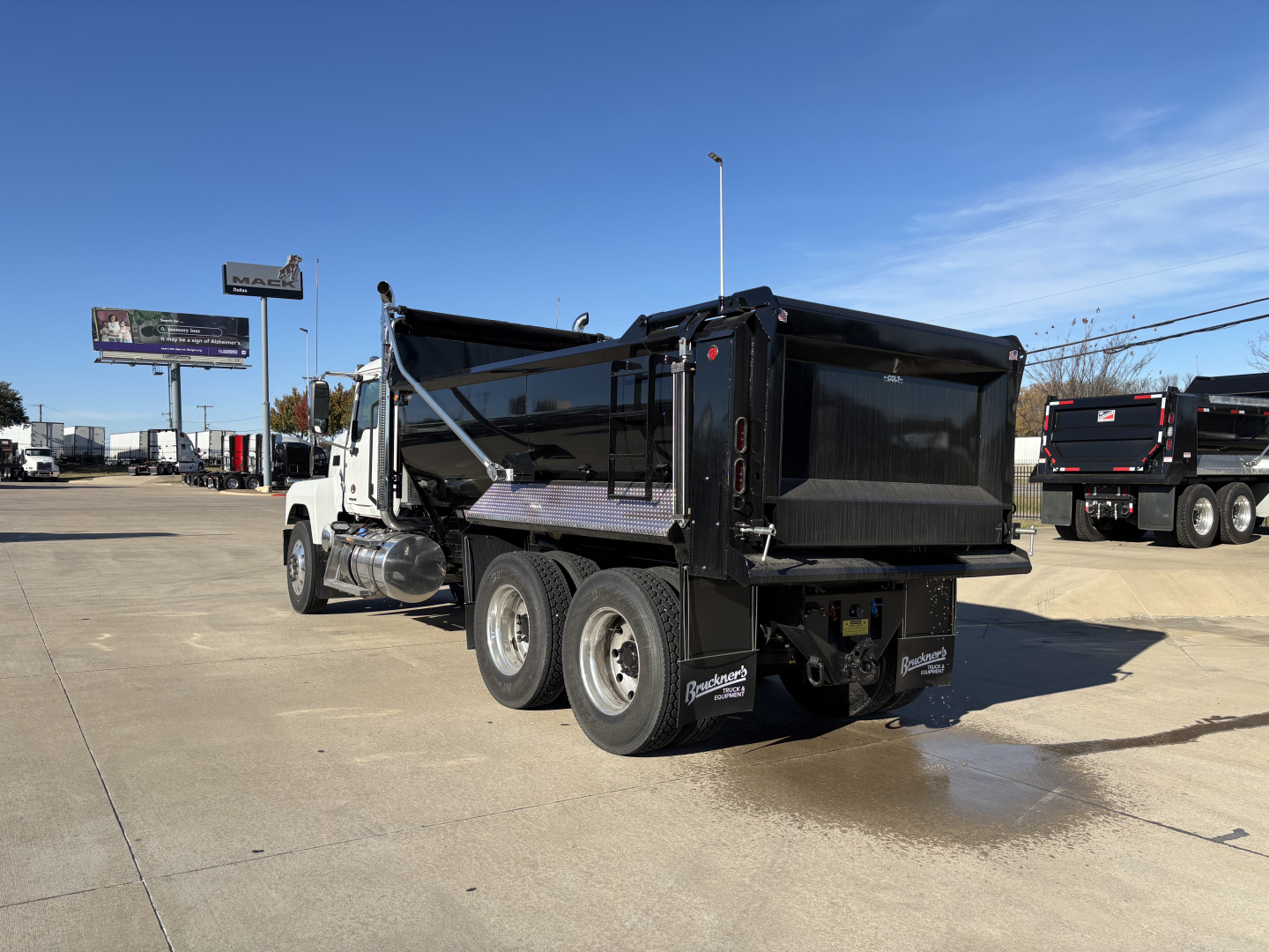 Dump truck demonstrating a Mack PI64 chassis with a black dump bed and white cab, side view showing triple rear axles and tailgate area.