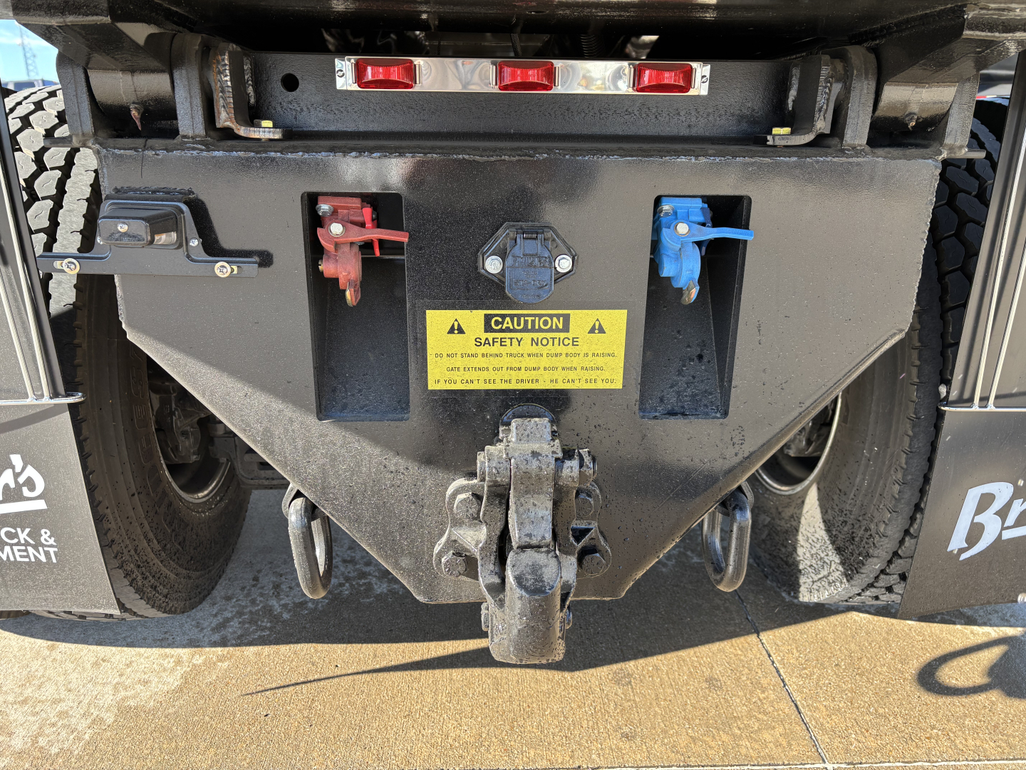 Mack PI64 rear view showing hitch, two color-coded battery disconnects (red and blue), and yellow caution label on dump truck bed.