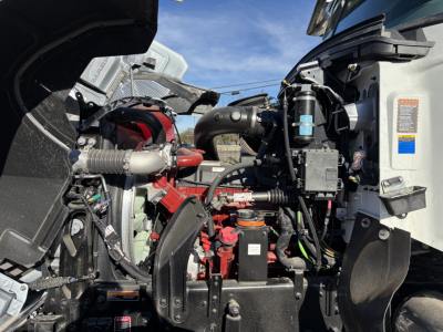 Engine bay of a 2026 Mack PI64 dump truck showing components like air intake, hoses, wiring, coolant reservoir, and battery near the engine block.