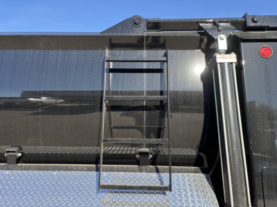 Dump truck bed with ladder/riser and hydraulic strut on right; black painted surface and diamond plate lower panel; small red indicator light near top edge.