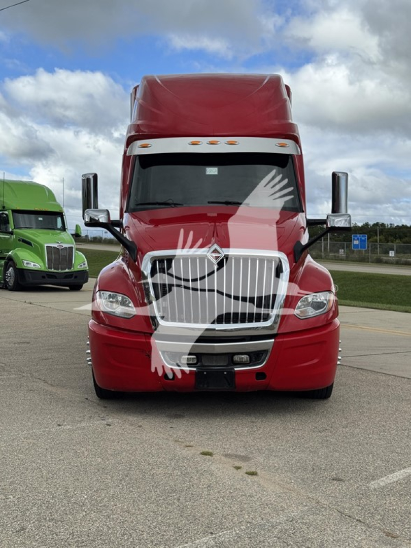 Front view of a red 2019 International LT semi-truck tractor in a parking lot, chrome grille with vertical bars, large windshield, sleeper cab, other green truck in background.