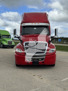 Front view of a red 2019 International LT semi-truck tractor in a parking lot, chrome grille with vertical bars, large windshield, sleeper cab, other green truck in background.