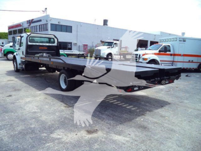 Flatbed tow chassis in a truck lot with Freightliner M2 106 in background and other vehicles.