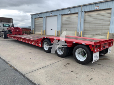 Lowboy trailer with red flatbed and dual rear axles, side rails and white wheels, parked outside a warehouse.