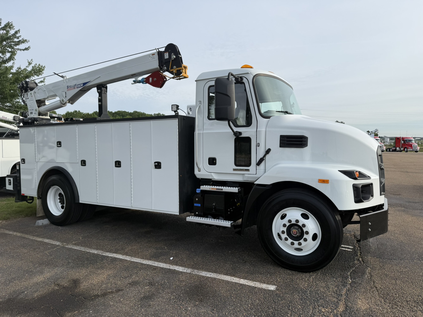 Mack MD6 service truck with crane arm, white utility compartments along the body, cab in view, side ladder steps, front bumper, and large front wheel in a parking lot.