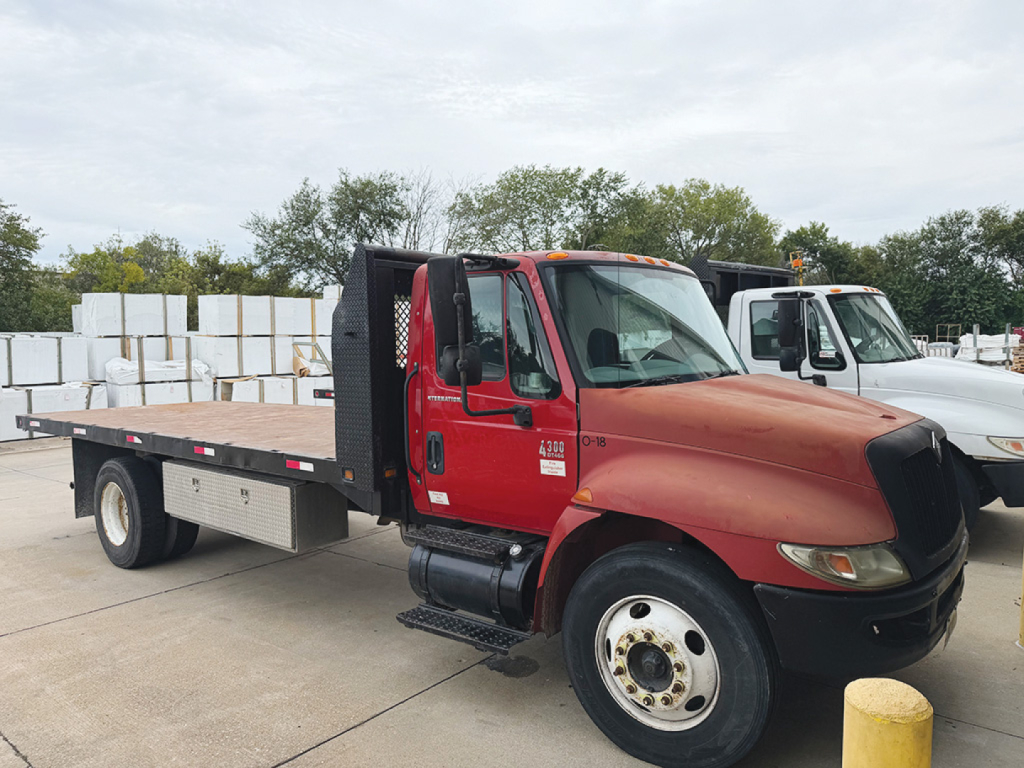 International 4300 conventional daycab truck with red cab and flatbed, side step, black grille, and storage boxes in bed; shows wheel, side mirror, and VIN tag area.