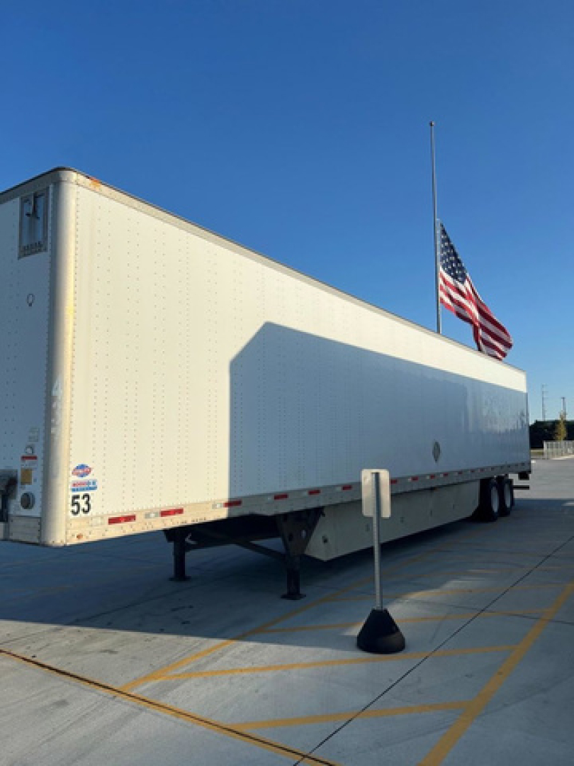 Utility van trailer showing side and rear with a flagpole and American flag, white exterior, trailer legs deployed, stock number 53 visible near rear left corner.