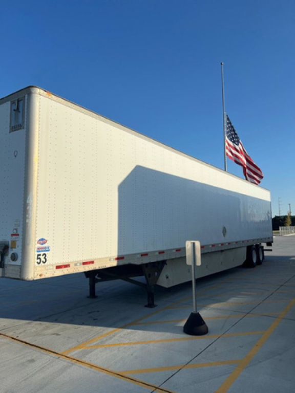 Utility van trailer showing side and rear with a flagpole and American flag, white exterior, trailer legs deployed, stock number 53 visible near rear left corner.