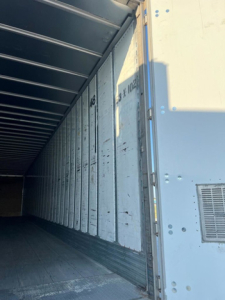 Interior of a trailer van showing corrugated metal walls with vertical seams, open left side; some faded markings on wall panels and a vent near the right edge.