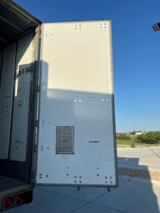 Rear exterior of a utility van trailer with white panels, side vent, and corner studs; visible loading area and open door on left; clear blue sky.