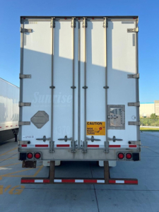 Rear view of a white utility van trailer with double doors, multiple latches, and safety decals visible on the back doors.