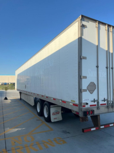 Trailer rear and side view showing enclosed box trailer with dual rear doors, white exterior, and tandem axle wheels.