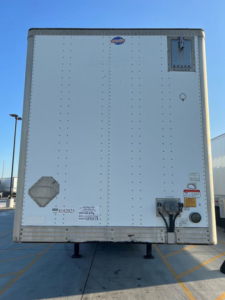 Rear view of a white utility trailer showing bumper area with access hatch, electrical plug, and stickers.