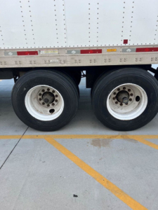 Trailer wheel area showing dual rear axles with white rims and black tires under a box trailer; part of the side with reflective tape and small stickers; concrete ground with parking lines visible.