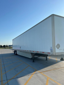 Utility van trailer in a parking lot, side view showing empty white box body with riveted panels and landing legs at front, blue sky above.