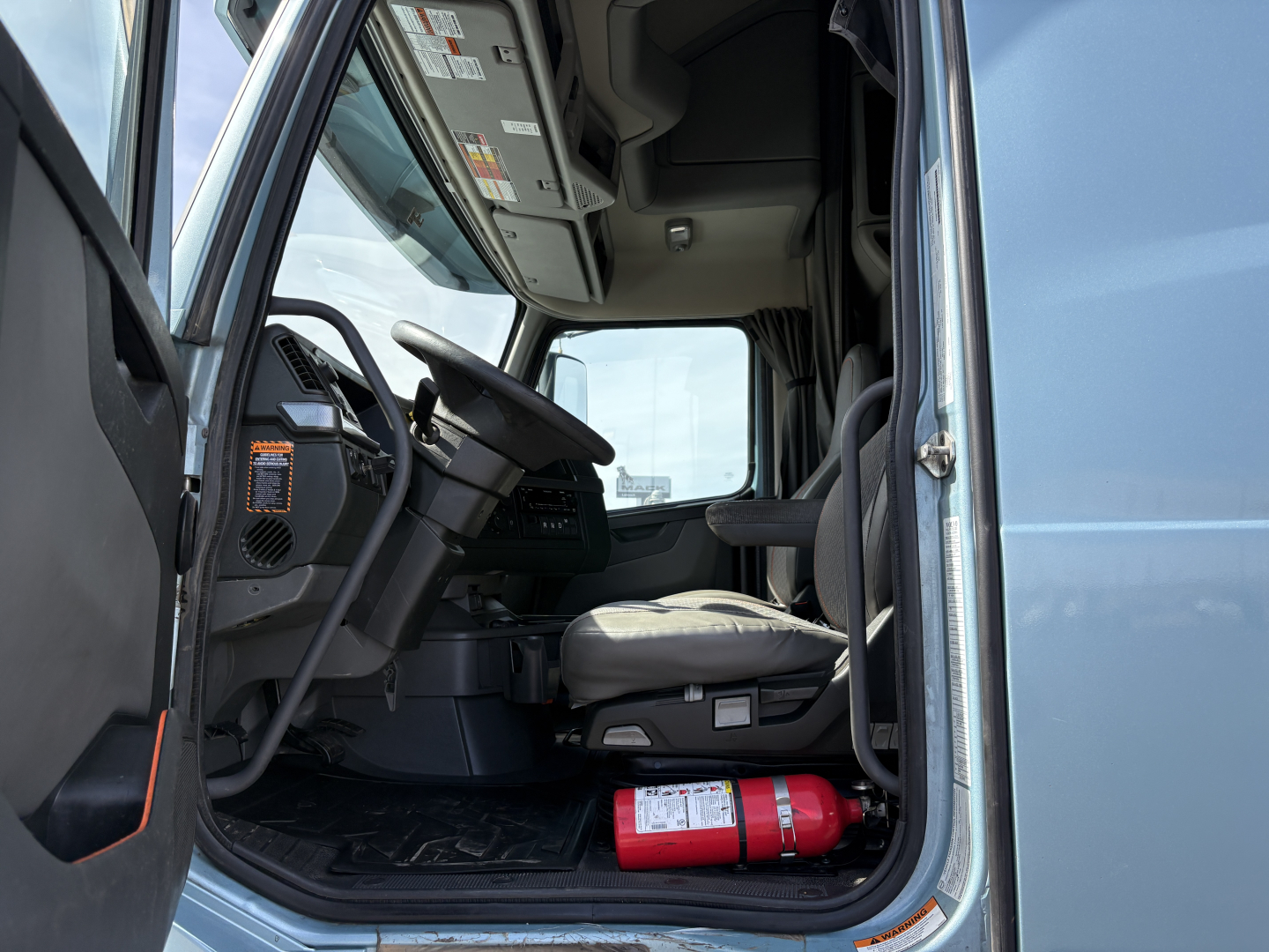 Volvo VNL64T760 truck cab interior view from open door showing steering wheel, dashboard, driver seat, and a fire extinguisher on the floor next to the pedals.