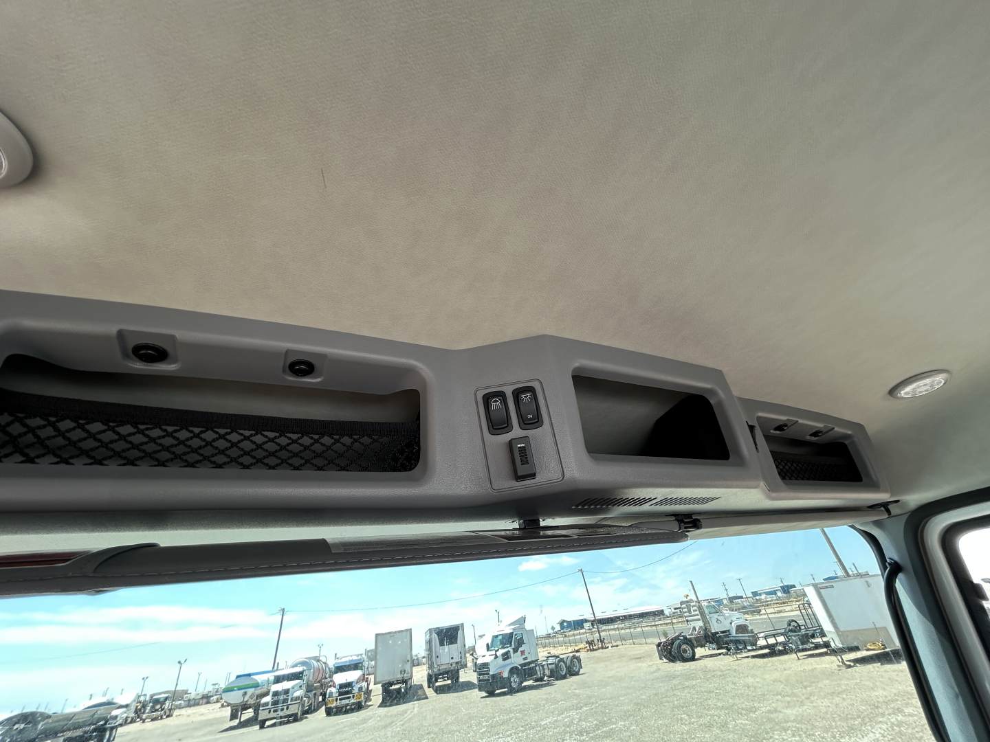Cab interior overhead console with storage compartments and switches, view through windshield to trucks and trailers in lot.
