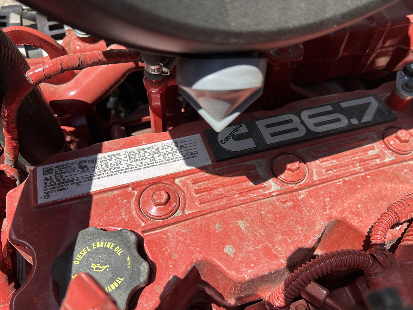 Engine bay of a new 2026 Mack MD7 showing the red engine cover with label and oil cap. Visible text includes 'B6.7' and various warning labels near the top.