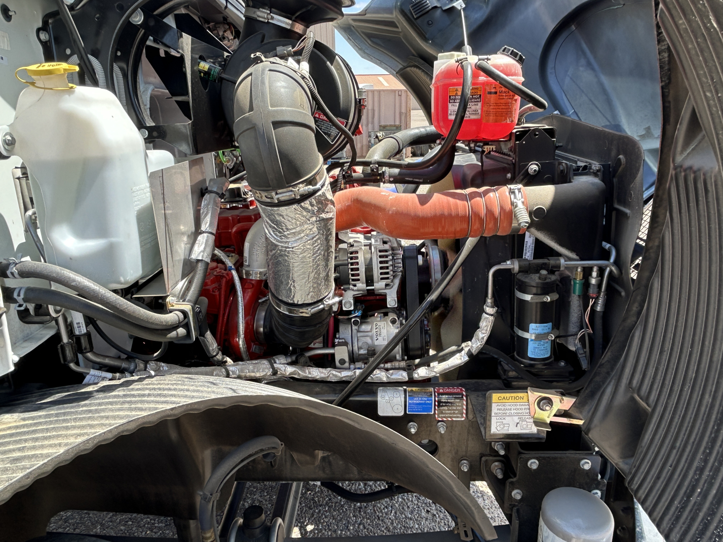 Engine bay of a 2026 Mack MD7 truck showing radiator coolant reservoir, air intake, hoses, alternator, and various engine components.