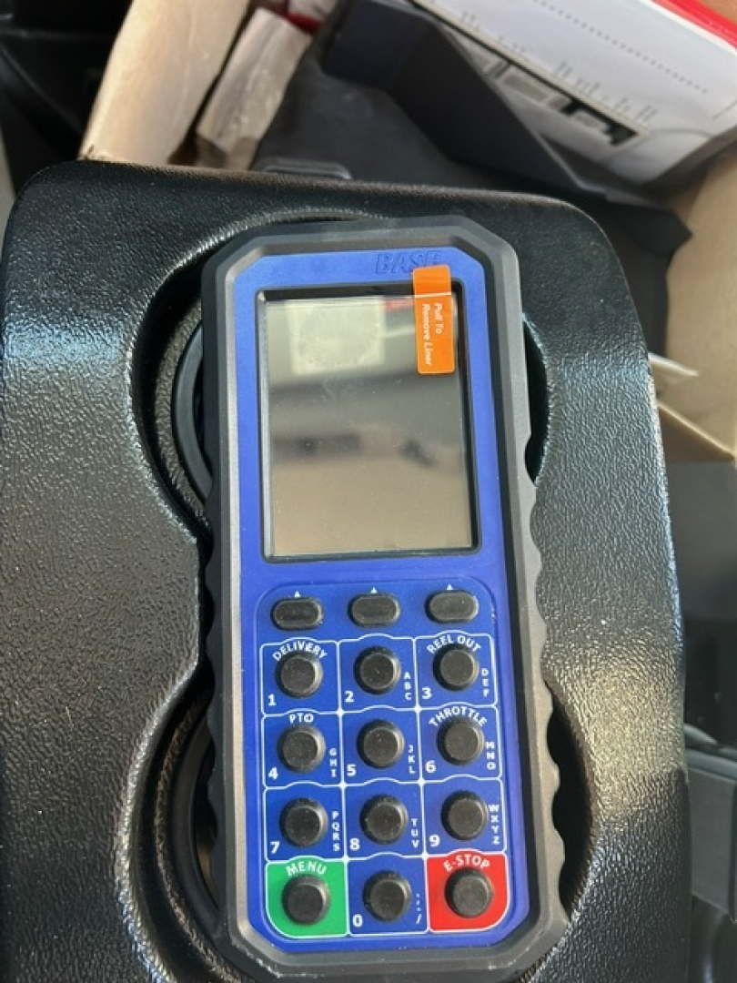 Blue handheld remote control device with numerous black buttons and small LCD screen; orange label at top-right corner; device sits in a vehicle compartment.